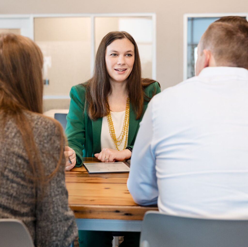 Business meeting: Woman in green blazer talks with clients at wooden table in bright office. Professional consulting services.