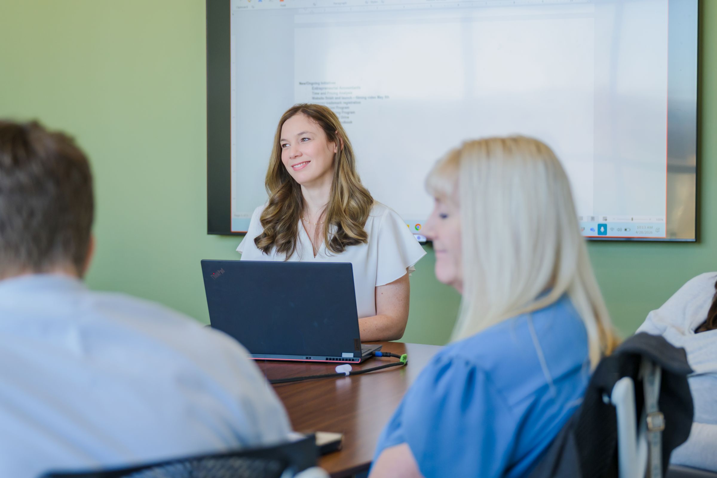 Business professionals meet in a conference room with a projector screen displaying notes in the background.